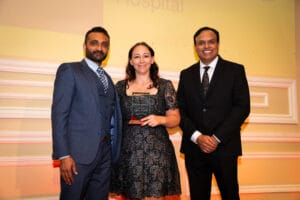 Three people in formal attire stand together indoors; the woman in the center holds a glass award and smiles at the camera.