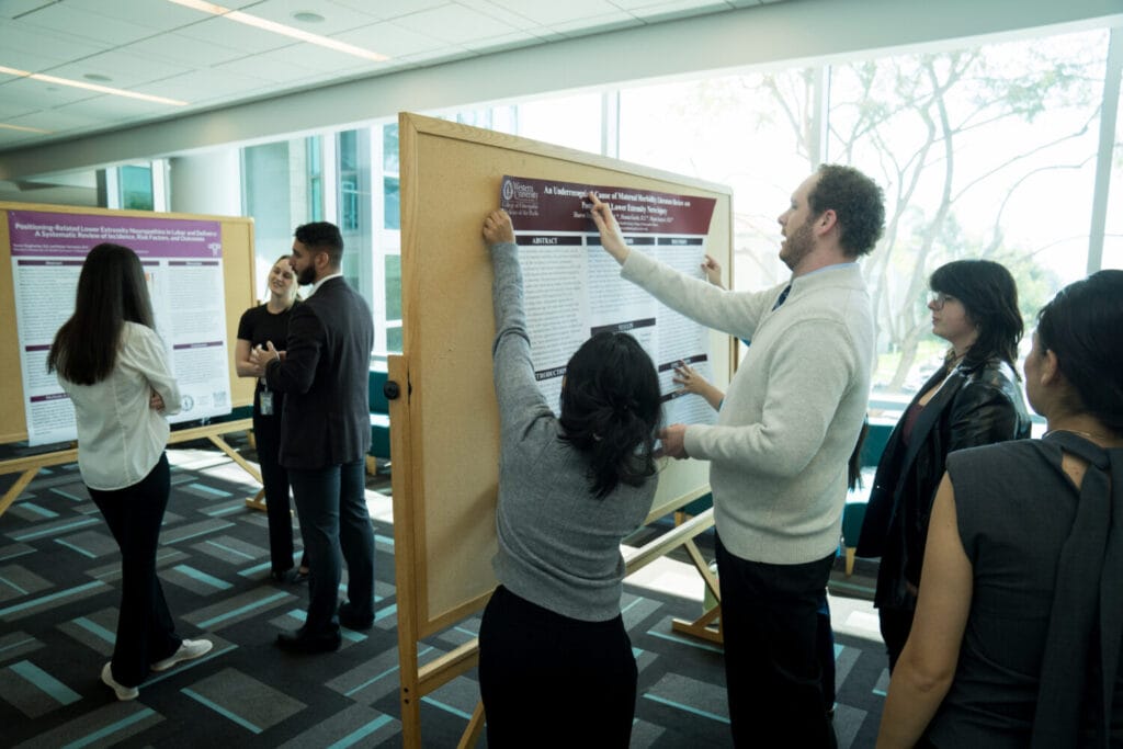 People stand and discuss research posters displayed on boards in a well-lit room with large windows.