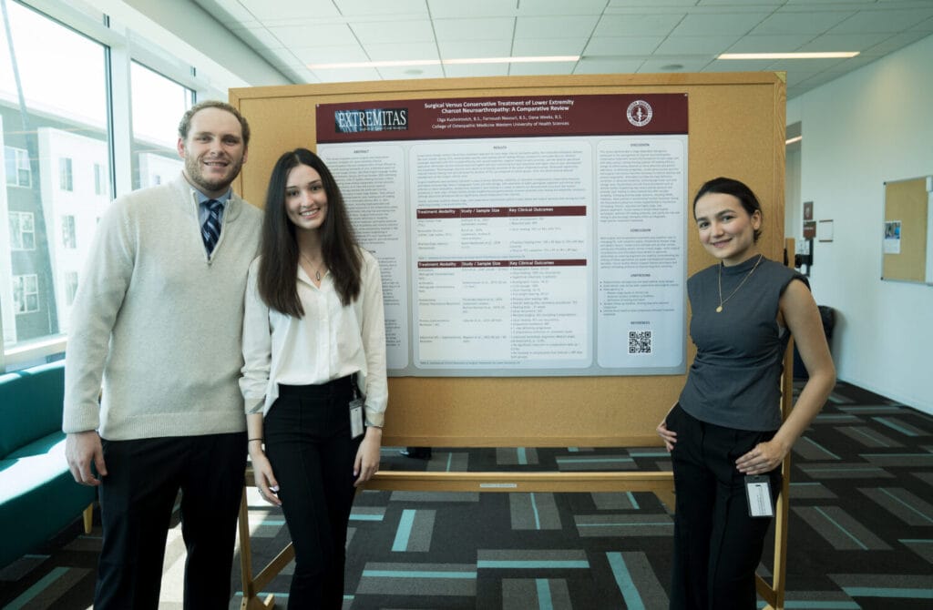 Three people stand in front of a research poster displayed on a bulletin board in a brightly lit room with windows and modern decor.