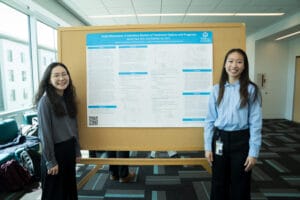 Two women stand on either side of a scientific research poster titled "Pediatric Melanoma: A Literature Review of Treatment Options and Prognosis" in a well-lit conference room.