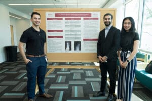 Three people stand in front of a scientific research poster on “Long-Distance Runners and Metatarsal Stress Fractures” in a modern indoor setting.