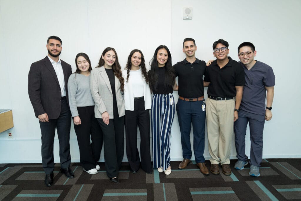 Eight people stand side by side in business casual attire, smiling at the camera in a brightly lit room with a geometric patterned floor.