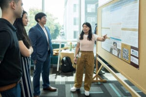 A woman presents research on a poster to three people in a brightly lit room with large windows and modern decor.