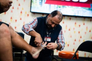 A man wraps athletic tape around another person's ankle in a brightly lit room, wearing a plaid shirt and a vest with an ID badge.