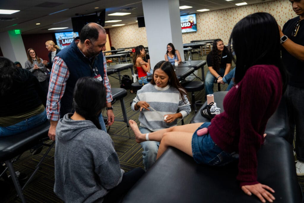 A group of people in a classroom setting practice bandaging techniques on a person's foot using medical tape, with others observing and talking in the background.