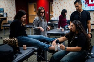 A woman wraps another woman's ankle with medical tape in a classroom, while two others observe and a man stands nearby.