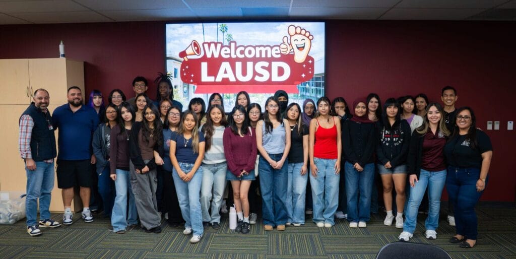 A group of students and adults pose together indoors in front of a screen displaying "Welcome LAUSD.