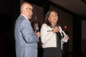 A man in a suit and a woman holding papers and a microphone stand on stage, speaking to an audience.