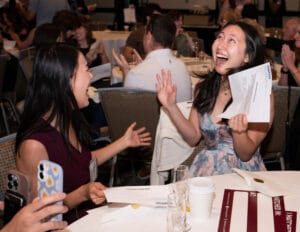 Two women sit at a table, smiling and laughing with excitement while holding envelopes at an indoor event. Other people and tables are visible in the background.