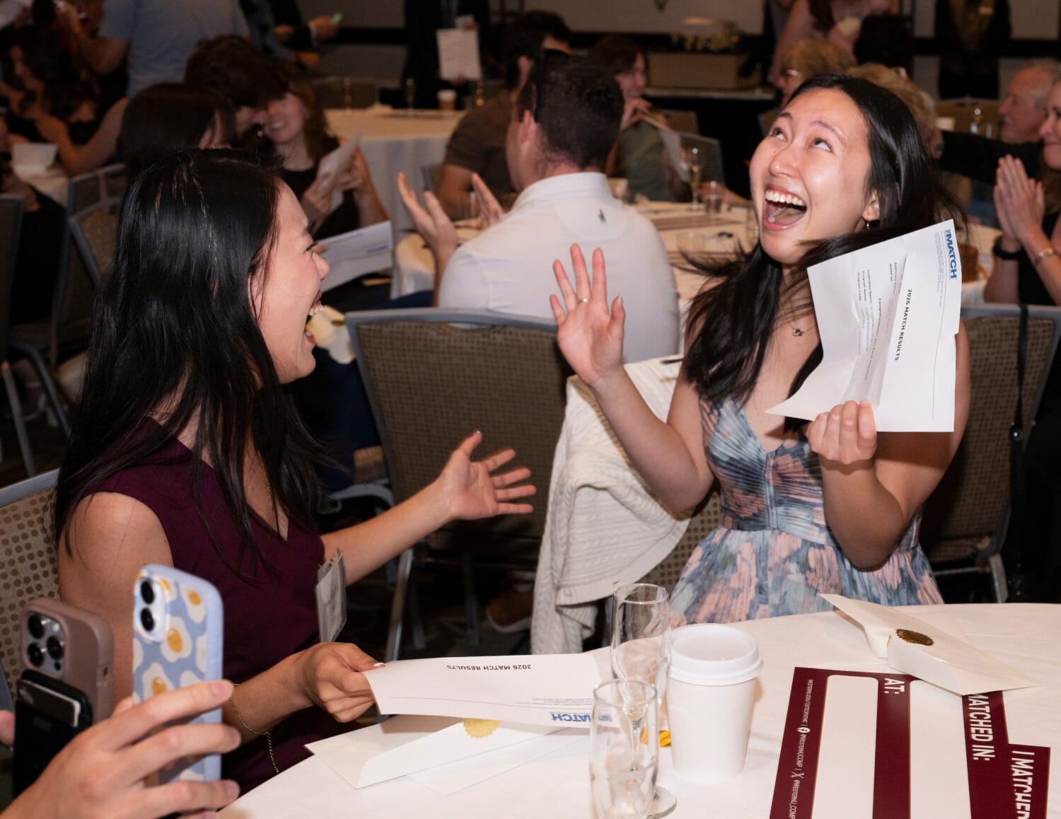 Two women sit at a table, smiling and laughing with excitement while holding envelopes at an indoor event. Other people and tables are visible in the background.