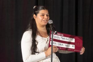 A woman stands at a microphone holding a sign that reads "Family Medicine" and "Loma Linda Uni" with a heart symbol, against a black background.