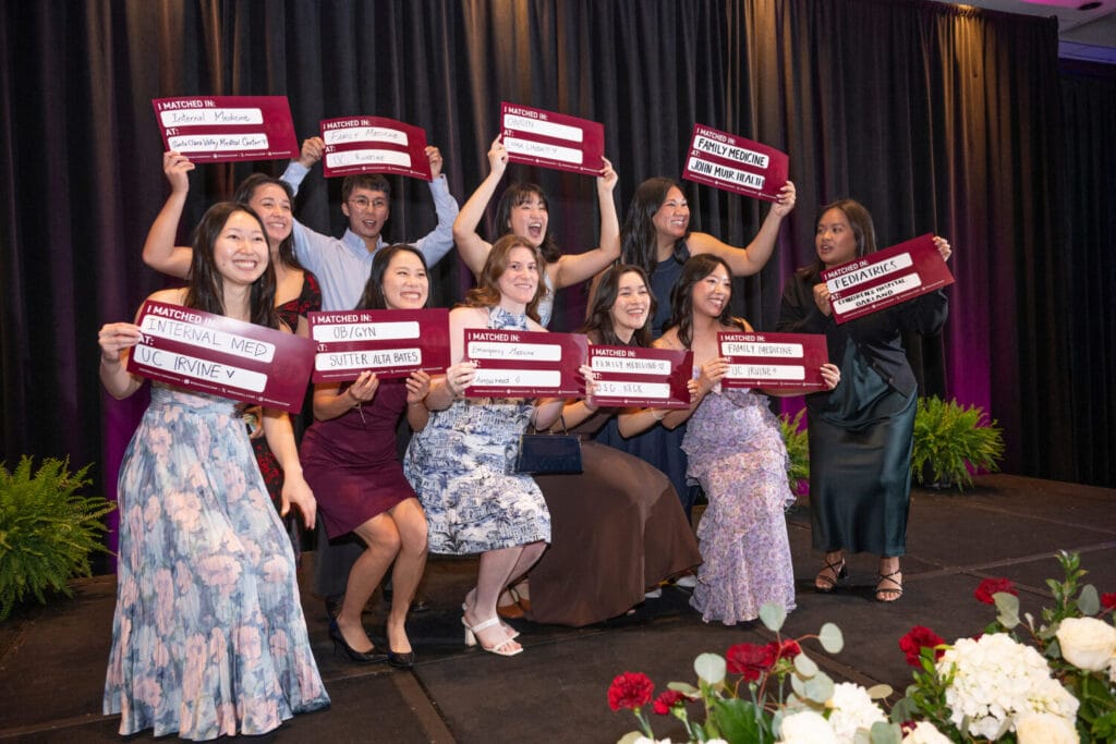A group of people on stage hold red signs with medical residency match results, smiling and posing for a photo at an event with black curtains and floral decorations.