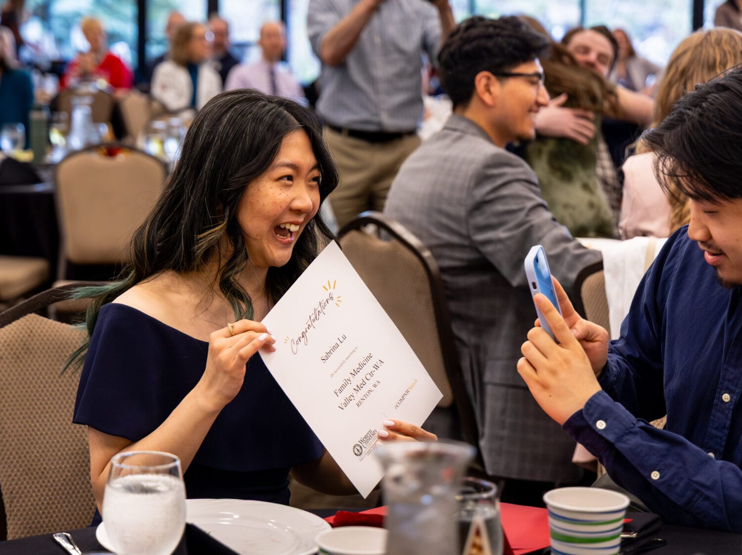 A woman smiles and holds up a certificate while a man takes her photo with a phone at a banquet event.