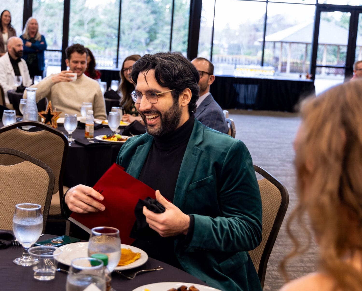A man in a green blazer sits at a round table, smiling and holding a red folder at an indoor event with several other seated guests.