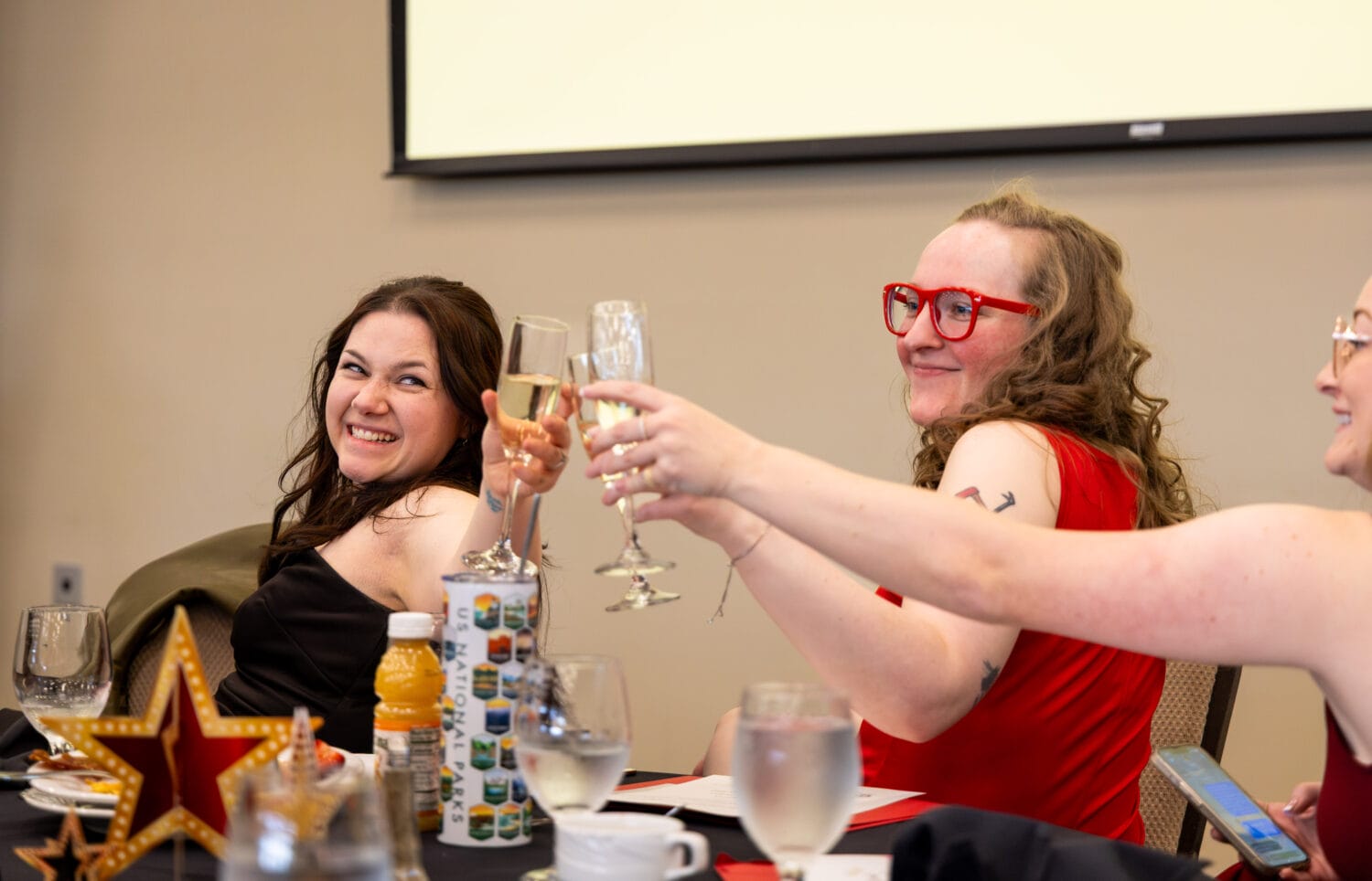 Three people sitting at a table raise and clink champagne glasses, smiling during an indoor celebration or event.