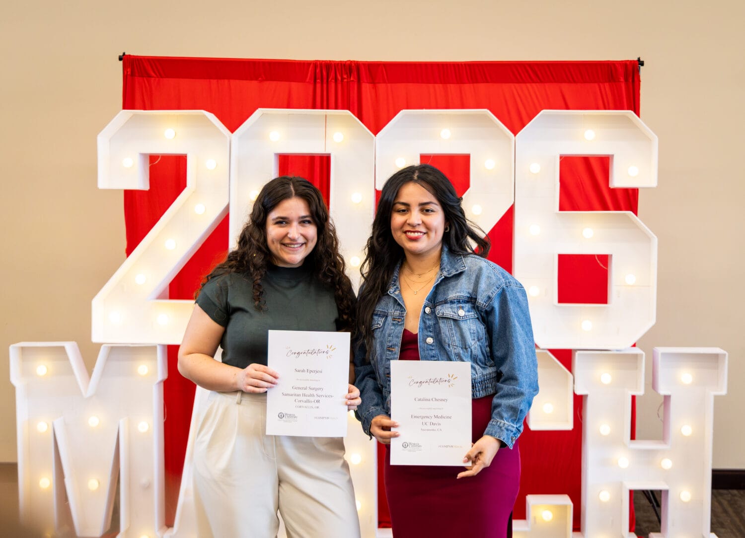 Two women stand in front of large lit "2026" numbers, each holding a certificate and smiling at the camera.