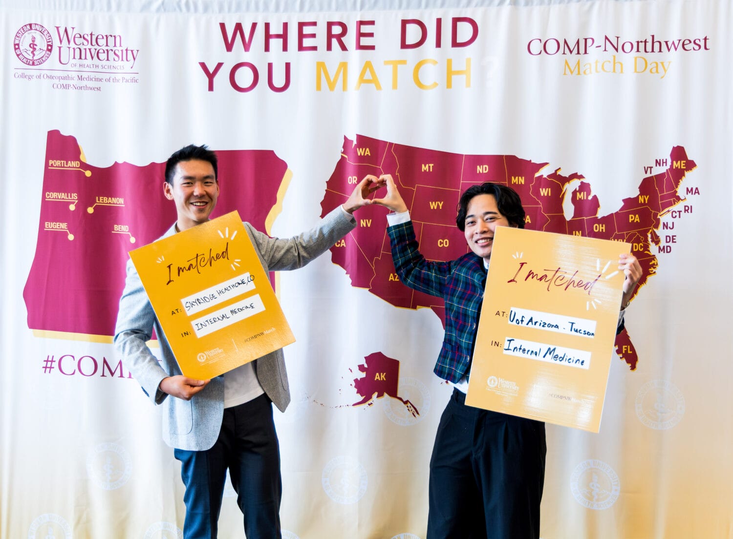 Two people stand smiling in front of a "Match Day" map backdrop, each holding yellow boards showing their medical residency matches. They form a heart shape with their hands.