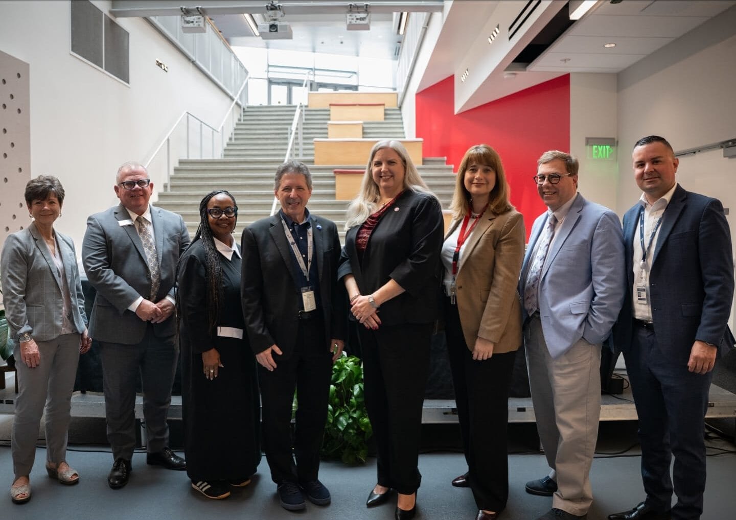 Eight professionally dressed people stand in a row indoors, in front of a staircase and a red accent wall.