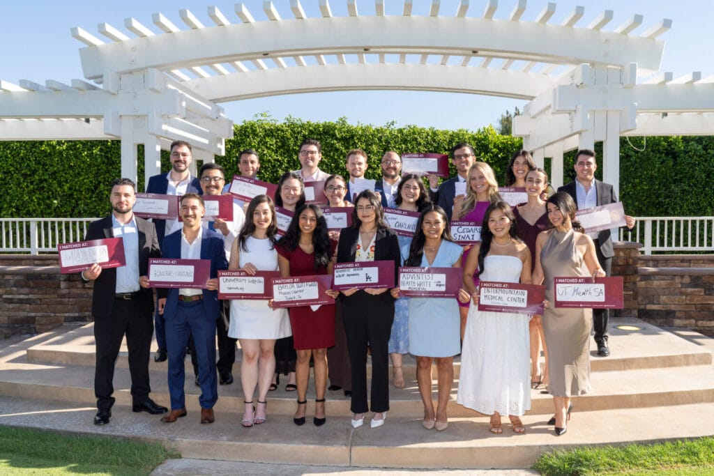 A group of adults dressed in business or semi-formal attire stand outdoors, each holding a sign with text, posing for a group photo under a white pergola.