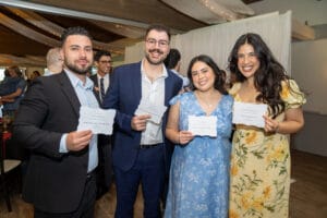 Four people in formal attire stand indoors, smiling and holding white cards with printed text, at what appears to be a social or celebratory event.
