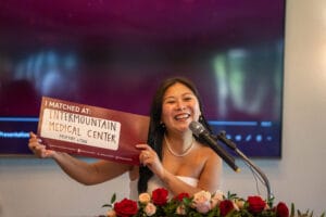 A woman stands at a podium with a bouquet of roses, smiling and holding a sign that reads, "I matched at: Intermountain Medical Center, Murray, Utah.