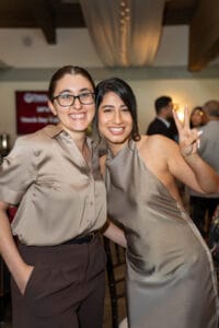 Two women pose together at an indoor event; one is smiling with her hand in her pocket, and the other is giving a peace sign while smiling towards the camera.