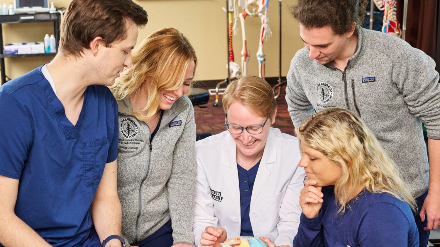Five people, including a person in a white coat, gather around a model of a human joint in a classroom, discussing or learning together.