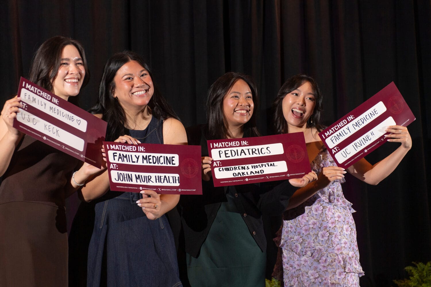 Four women smile and hold signs showing their medical residency match results in family medicine and pediatrics at various institutions, standing in front of a dark curtain.