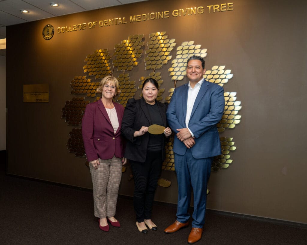 Three people stand in front of a wall display reading "College of Dental Medicine Giving Tree"; one person holds a gold plaque.