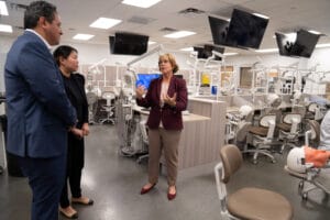 Three people stand and talk in a modern dental training lab filled with dental chairs, equipment, and monitors.