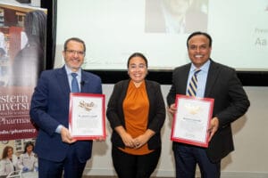 Three people pose indoors; two men in suits hold framed award certificates while a woman in business attire stands between them, smiling. A presentation screen and university banner are visible behind them.