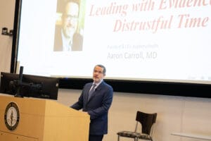 A man in a suit stands at a podium giving a presentation titled "Leading with Evidence in a Distrustful Time" with Aaron Carroll, MD, displayed on the screen behind him.