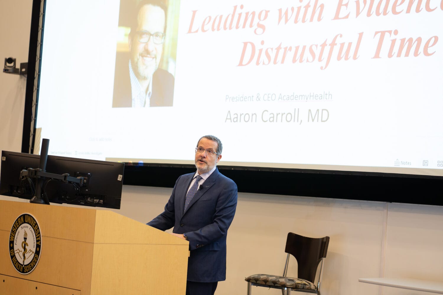 A man in a suit stands at a podium giving a presentation titled 