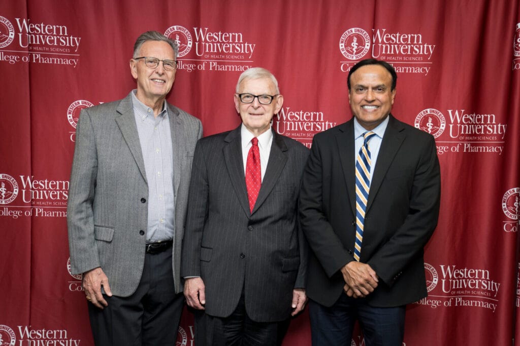 Three men in business attire stand in front of a Western University of Health Sciences College of Pharmacy backdrop.