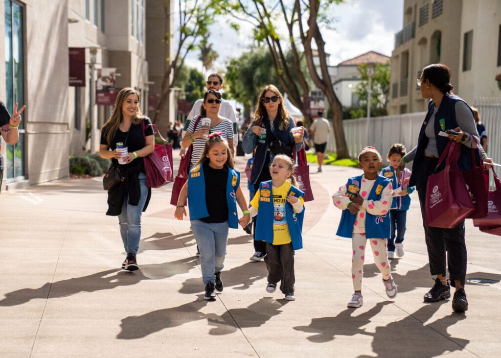 A group of adults and children wearing matching blue vests walk together on a sunny sidewalk, carrying bags and holding snacks.