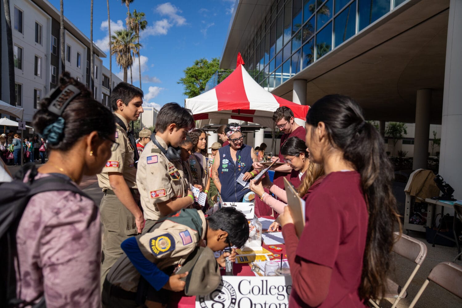 A group of scouts and students gather at an outdoor information booth under a red and white tent, with buildings and palm trees visible in the background.