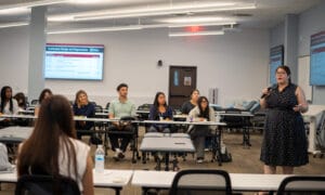 A woman in a black dress speaks into a microphone while seated students listen in a classroom with presentation slides displayed on screens.