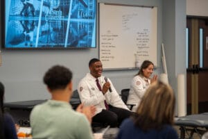 A man and a woman in white coats speak and interact with an audience in a classroom; a presentation screen displays "Western University" in the background.