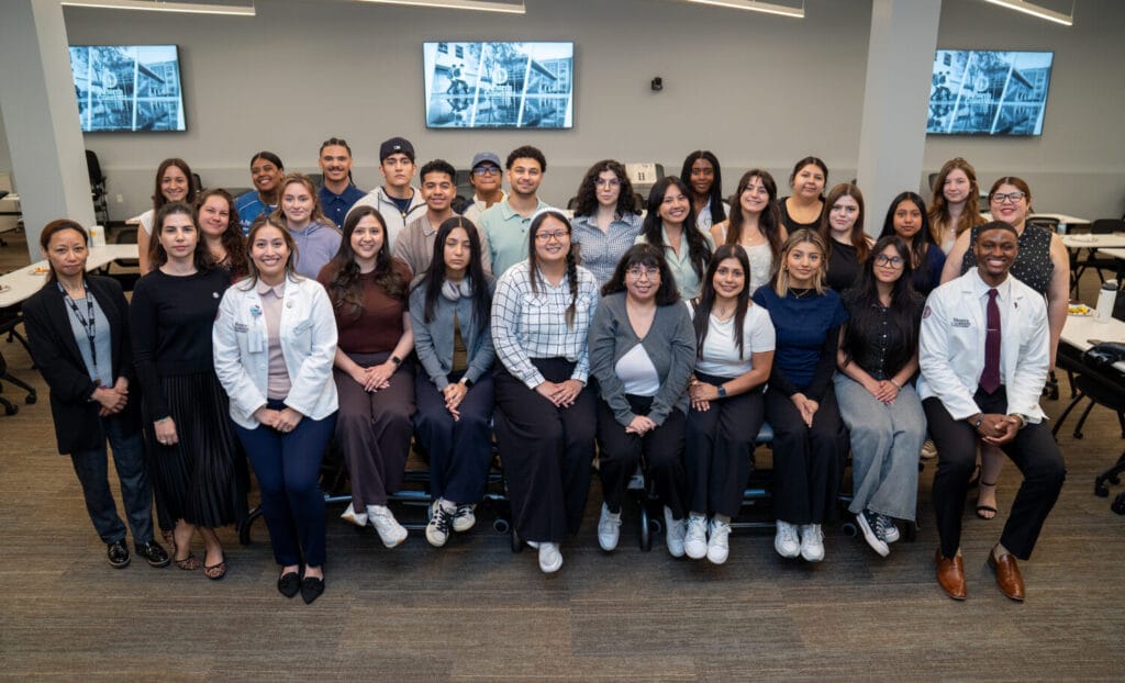A group of students and two people in white coats pose together in a classroom with large tables, chairs, and screens on the walls in the background.