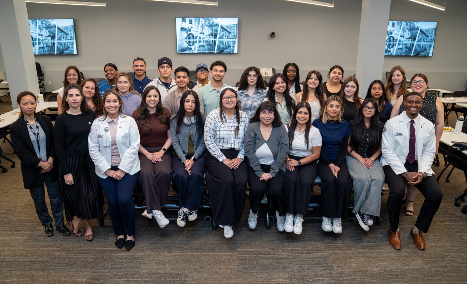 A group of students and two people in white coats pose together in a classroom with large tables, chairs, and screens on the walls in the background.