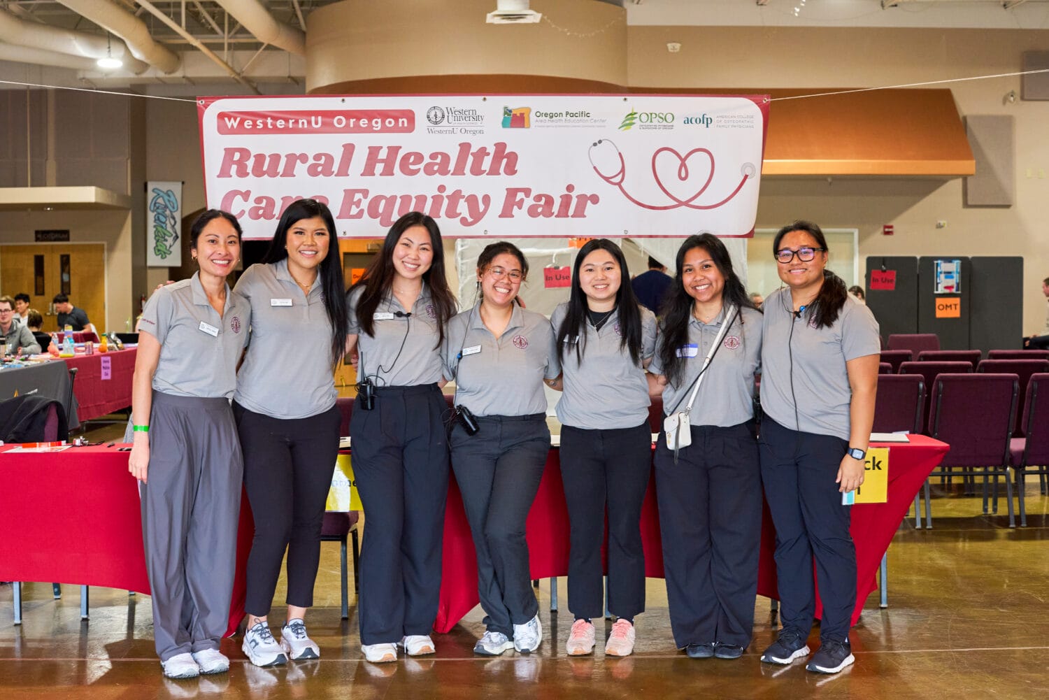 Eight people in matching gray shirts stand and smile in front of a red table and banner reading “Rural Health Care Equity Fair” at an indoor event.