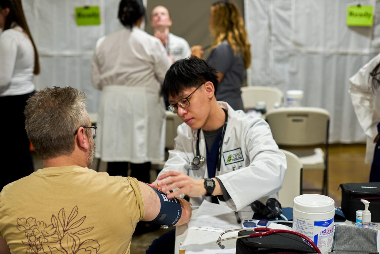 A healthcare professional checks a patient's blood pressure at a clinic or medical event, with other staff and patients visible in the background.