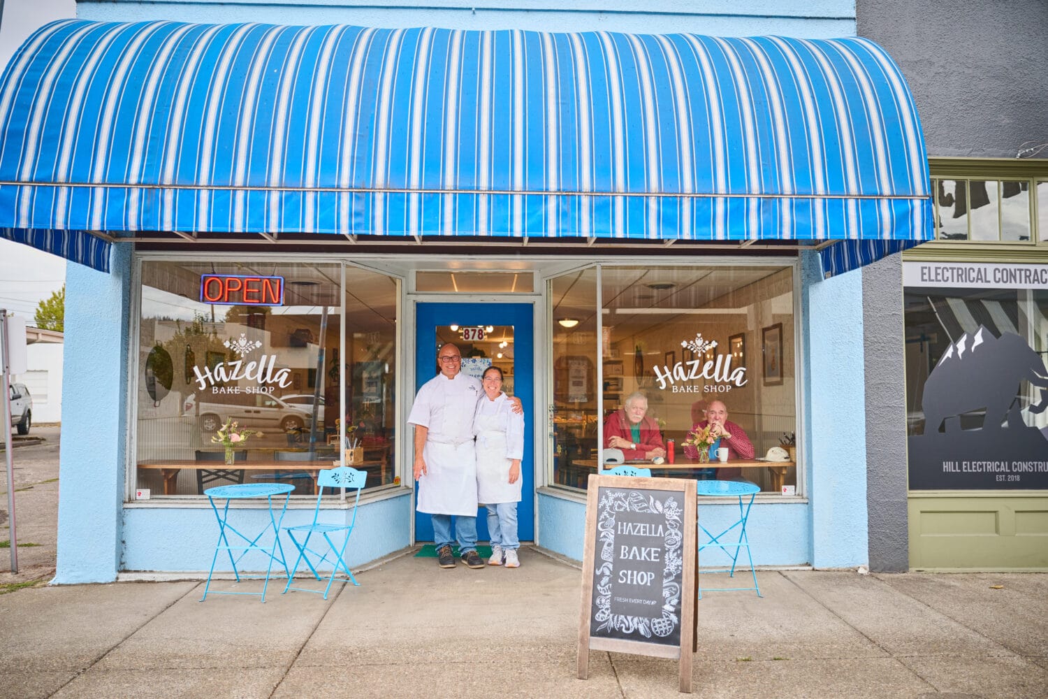 Two people in white aprons stand outside Hazella Bake Shop, which has a blue awning, outdoor seating, and an open sign. A chalkboard sign is in front of the bakery.
