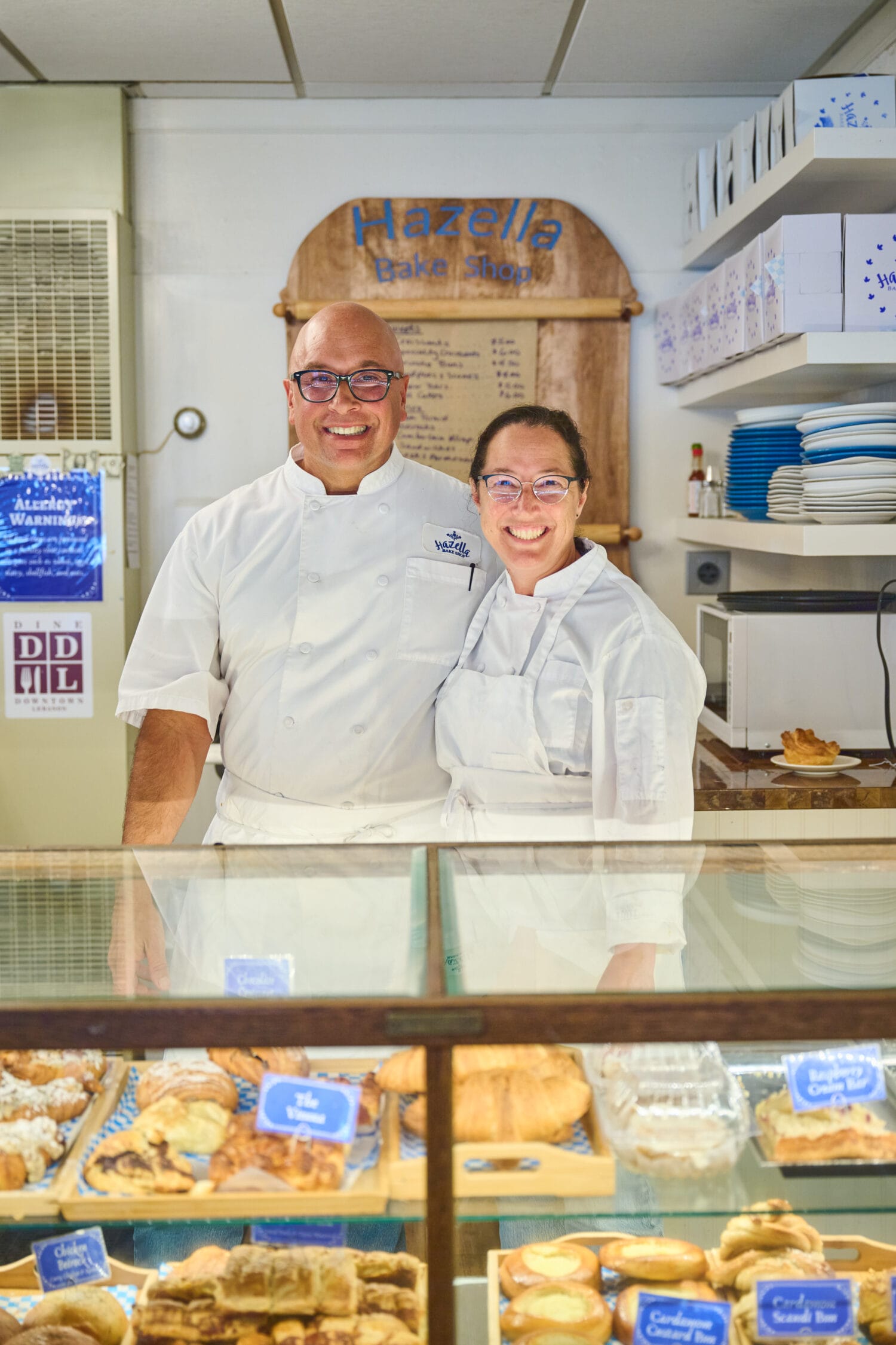 Two bakers in white uniforms stand smiling behind a glass display case filled with assorted pastries in a bakery named "Hazella Bake Shoppe.