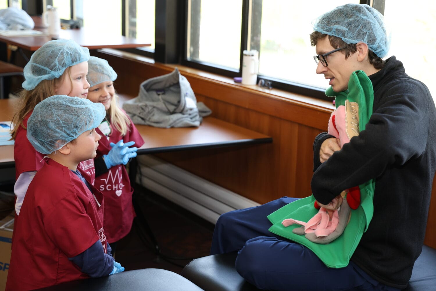 An adult wearing a hairnet shows three children a fabric model of human organs in a classroom setting. All are wearing hairnets and the kids are in matching maroon shirts.