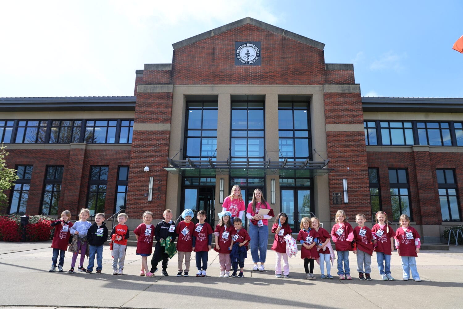 A group of young children and two adults stand in a line outside a large brick school building on a sunny day.