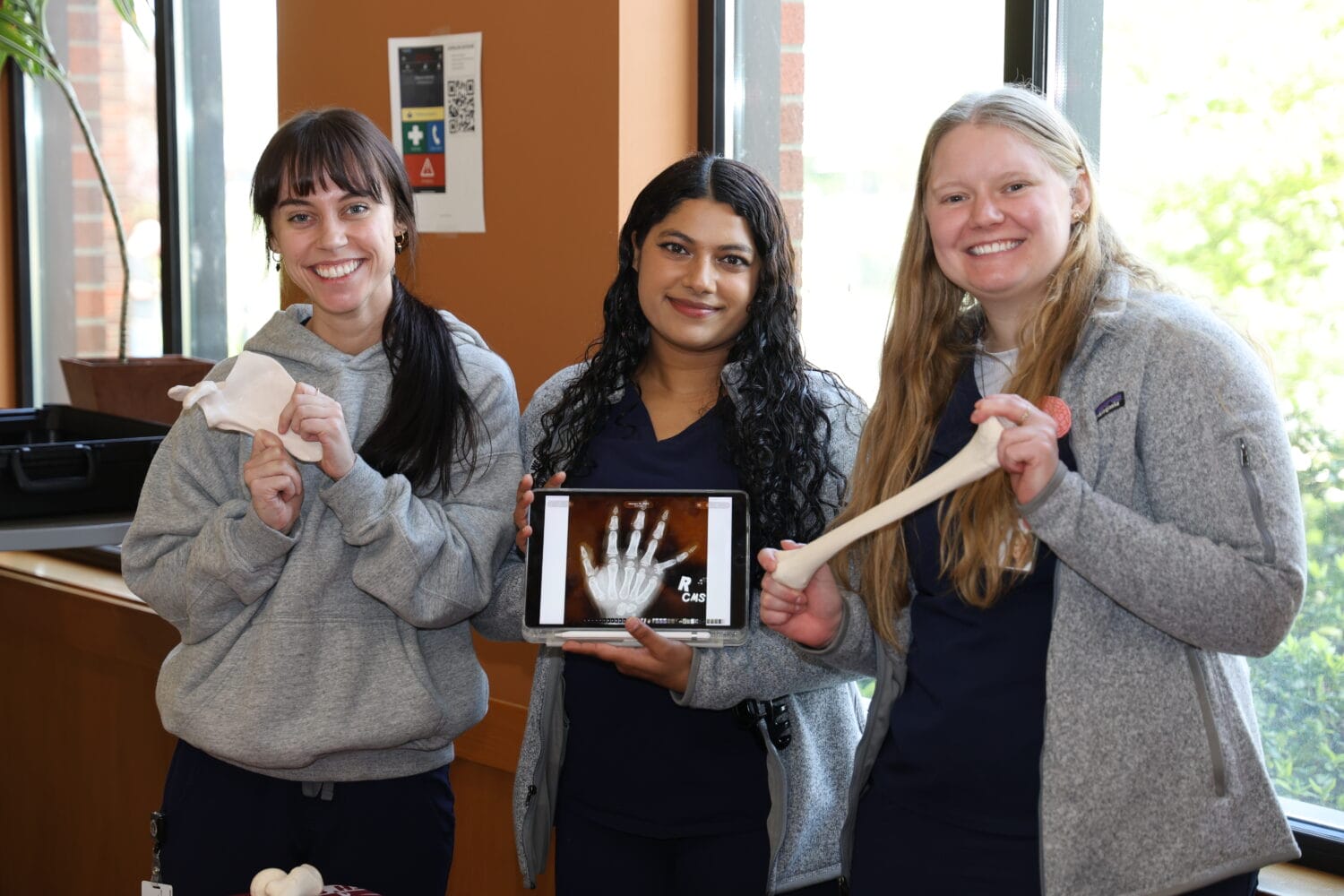 Three women in scrubs stand indoors; one holds a bone model, another displays a hand X-ray on a tablet, and the third holds a long bone model.
