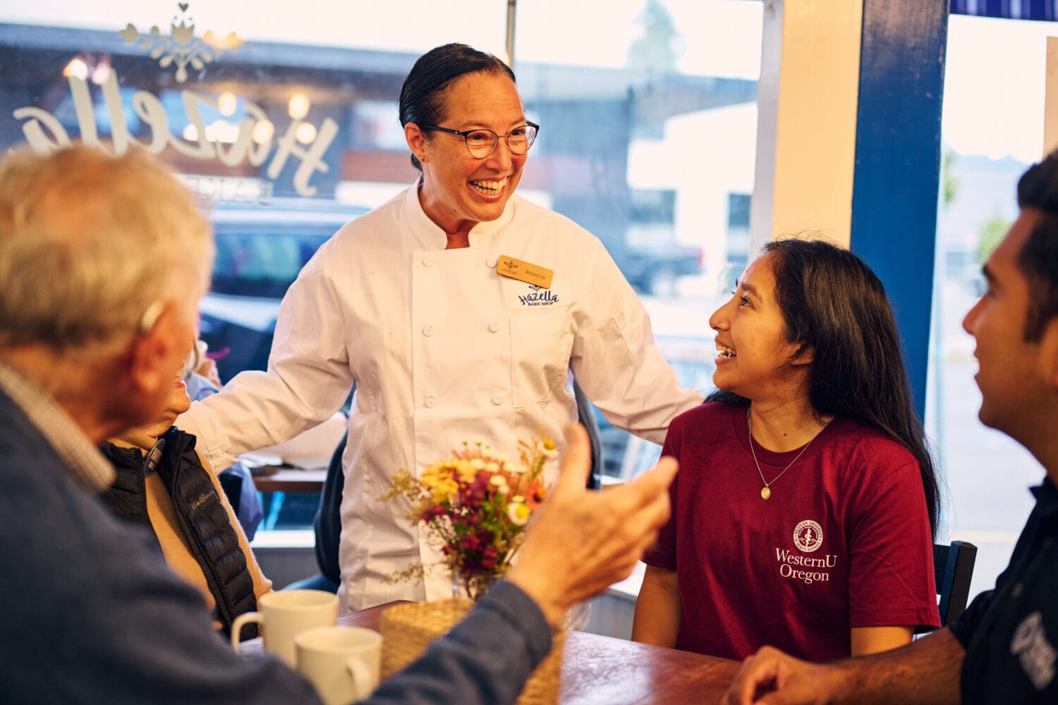 A chef in a white coat smiles and talks with three seated customers at a table in a café.