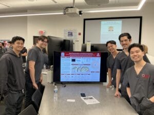 Six people in scrubs stand around a table, smiling, with a scientific poster displayed on a monitor in a classroom or conference setting.