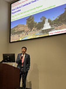 A man in a suit speaks at a podium in front of a projected slide titled "Stem Cells in Craniofacial Tissue Regeneration" at a scientific presentation.